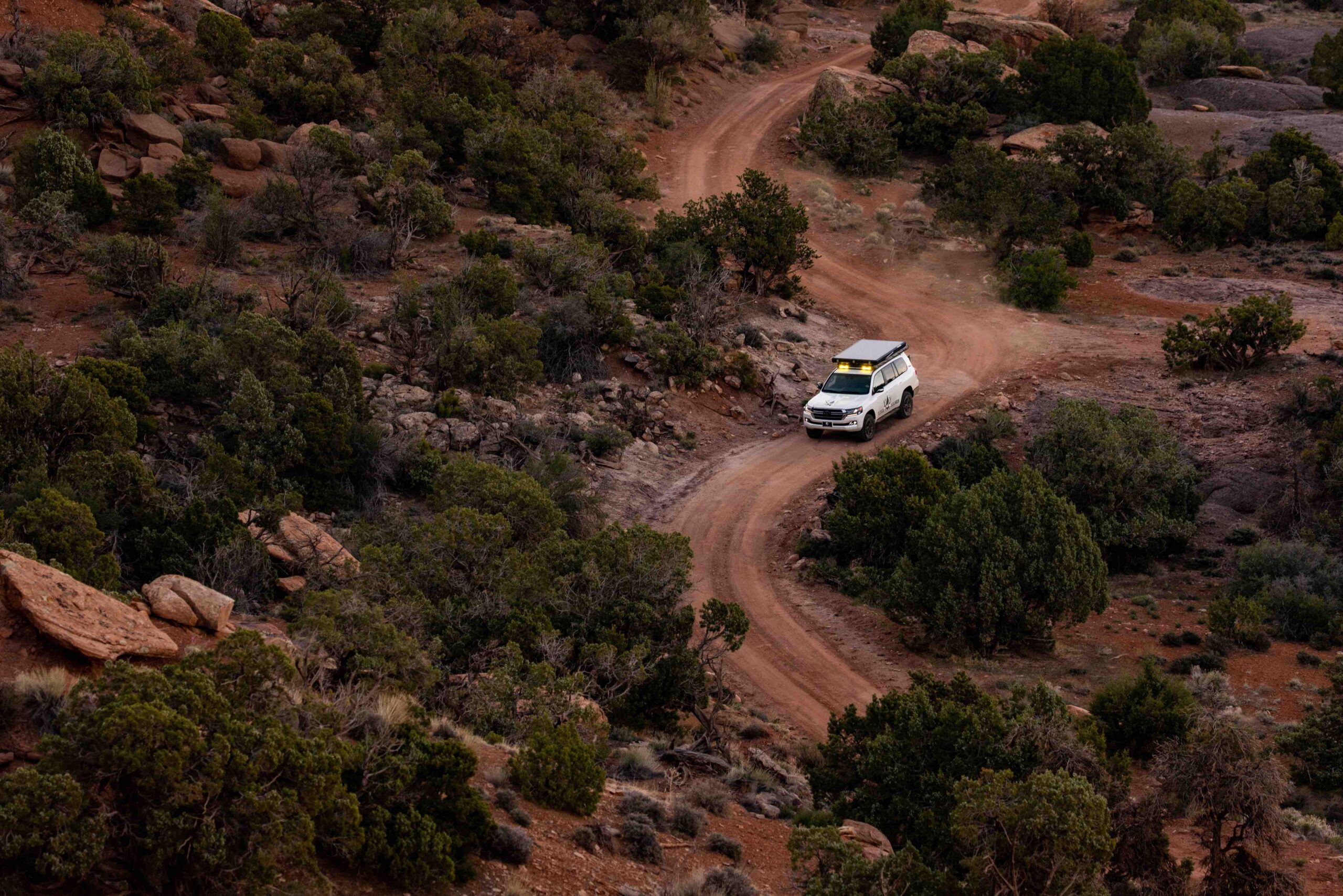 Captured from above, this aerial photo showcases the Colorado Overlander's Land Cruiser confidently navigating a vibrant red dirt road. Surrounded by piñon trees, the dust clouds billow up from behind the tires, accentuating the adventurous spirit of the journey. The dynamic scene captures the essence of off-road exploration and the rugged beauty of the terrain beneath the Land Cruiser's wheels.