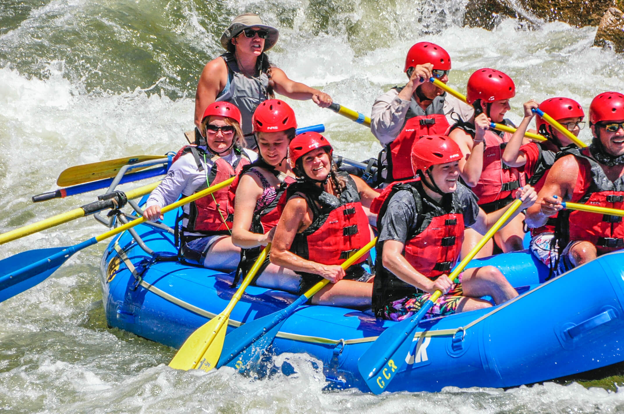 An image of rafters and their guide charging through river rapids. It is apparent that the guide is giving paddle instructions to the group.