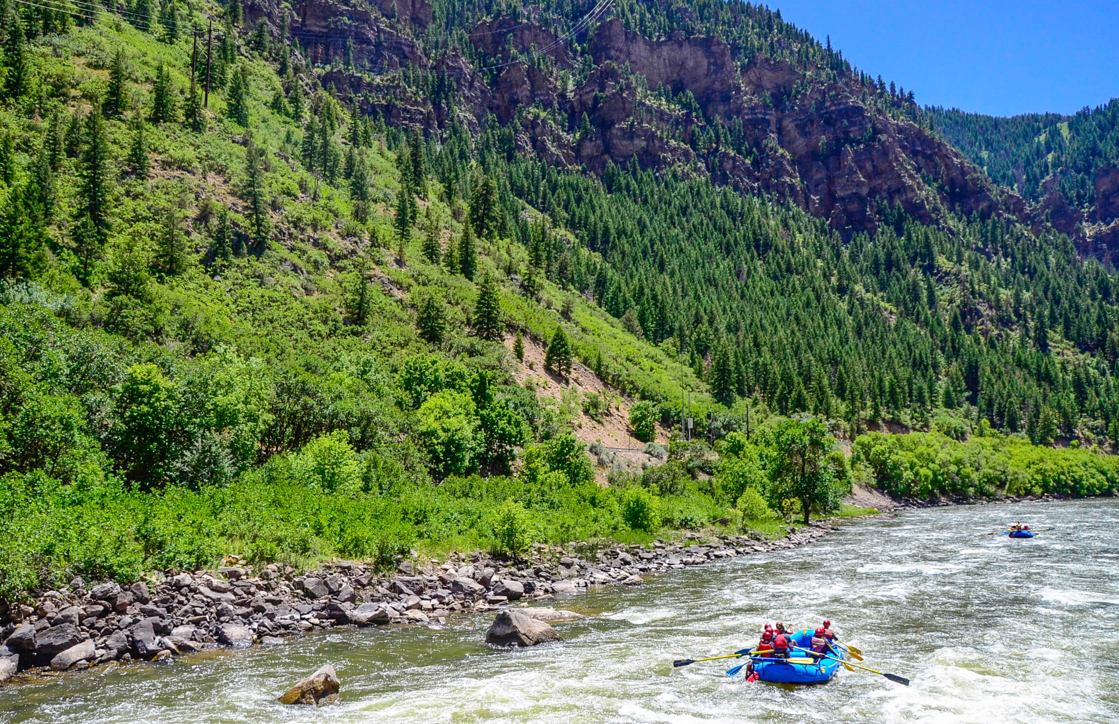 An image of a boat of rafters on the Colorado River with the beautiful green trees and rocky mountainside of Glenwood Canyon in the background.