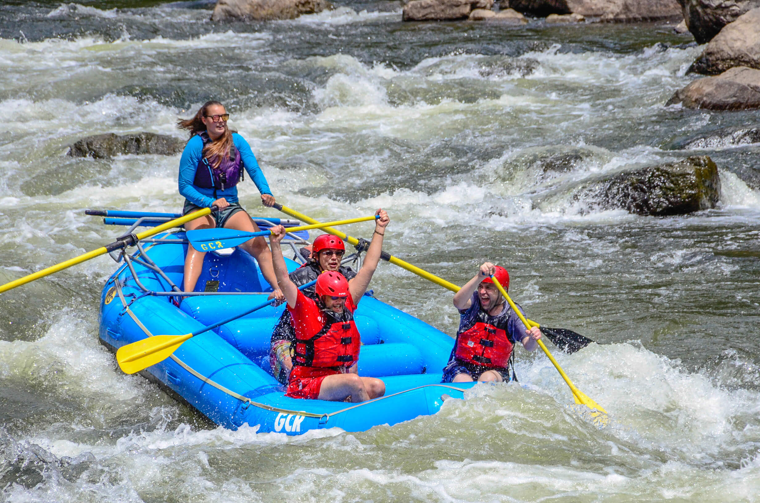 An image of a group of three rafters and their guide paddling through rapids. One rafter lifts paddle over his head in victory and celebration of the activity.