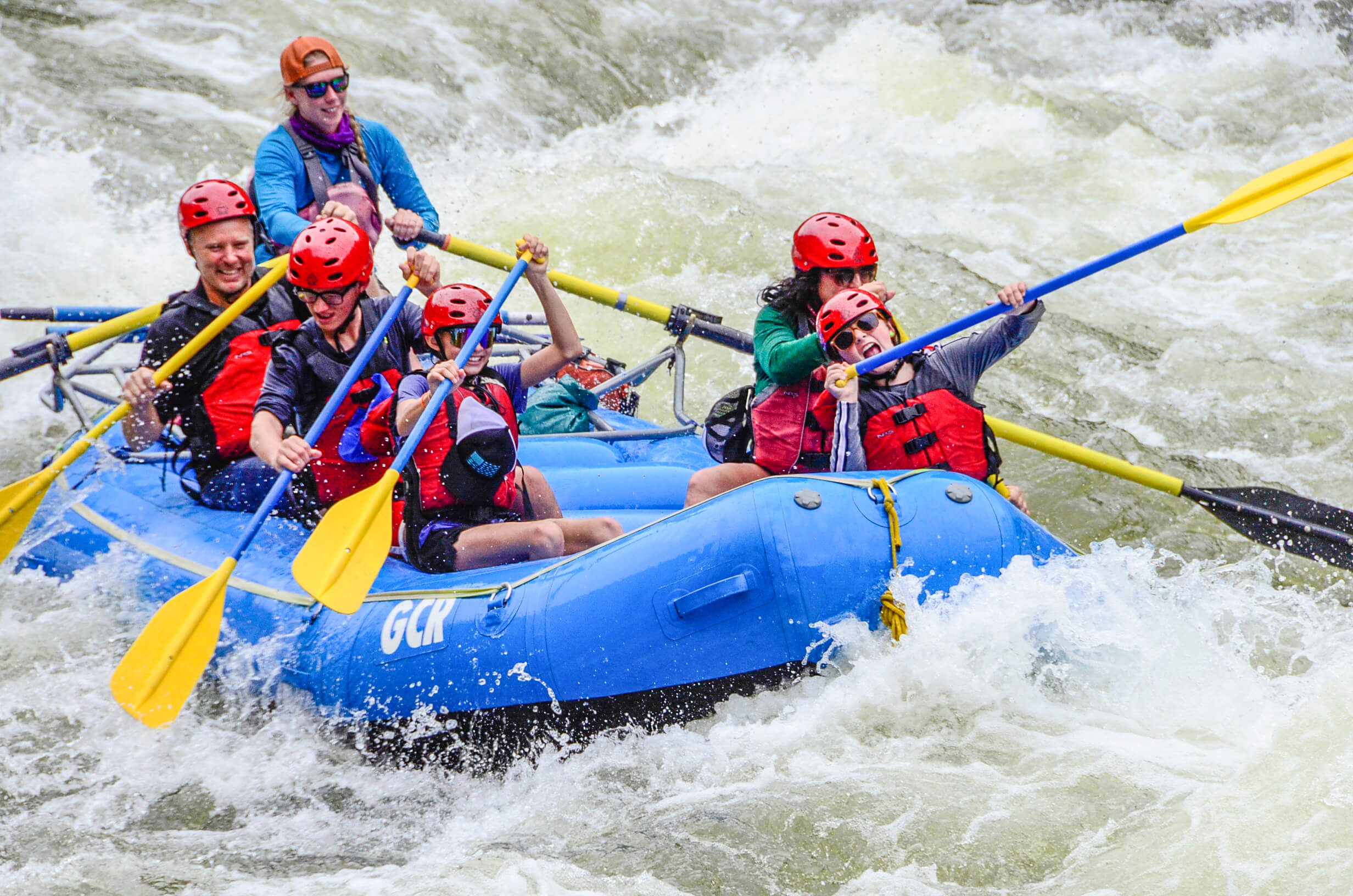 Image of a family with two young girls paddle through a set of rapids- the girls express excitement and joy as they celebrate with their smiles.